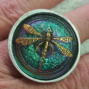 May include: A silver ring with a round, iridescent green, blue, and purple glass cabochon featuring a gold dragonfly design.