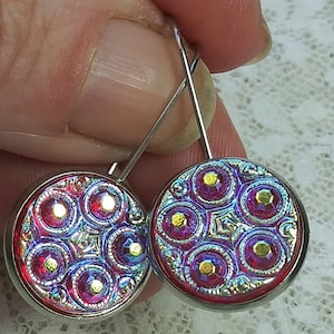 May include: A pair of silver dangle earrings with iridescent pink and purple glass cabochons. The cabochons have a geometric pattern with a silver metallic finish.