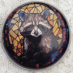 May include: A circular pin featuring a detailed illustration of a raccoon with black and white facial markings. The raccoon is set against a background of multicolored geometric shapes resembling stained glass in shades of yellow, orange, and blue.