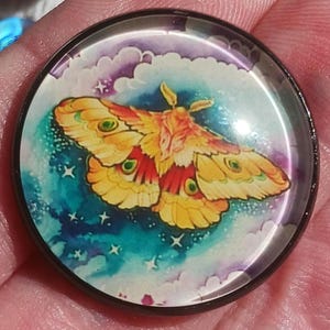 May include: A round, glass-covered button featuring a colorful moth illustration. The moth has yellow, orange, and red wings with green eye-like markings. The background is a mix of blue, purple, and white, with stars and clouds.