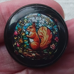 May include: A round, black button featuring a stained-glass design with a red squirrel. The squirrel is surrounded by colorful foliage and flowers, set against a blue and white background. The button has a glossy finish.