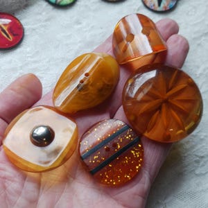 May include: A collection of orange and amber-colored buttons in various shapes and designs. Some buttons have a glossy finish, while others feature carved patterns or decorative elements. The buttons are made of a translucent material and have buttonholes.