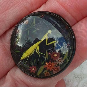 May include: A round, black button with a detailed illustration of a yellow praying mantis. The mantis is set against a dark background with colorful flowers and foliage. The button is held in a hand.