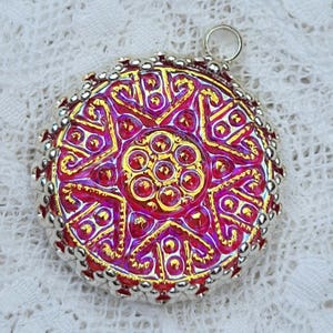 May include: A circular pendant featuring a vibrant magenta and gold star-shaped pattern with raised metallic detailing. The center is adorned with a cluster of small gold circles, and the outer edge is finished with a silver beaded border.