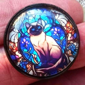 May include: A round, black-rimmed pendant featuring a Siamese cat illustration. The cat has blue eyes and is set against a stained-glass style background with blue, purple, and orange hues. The design is detailed and artistic.