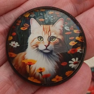 May include: A round, decorative pendant featuring a detailed illustration of a fluffy orange and white cat amidst a field of colorful flowers. The cat has green eyes and a serene expression. The pendant has a dark, metallic border.