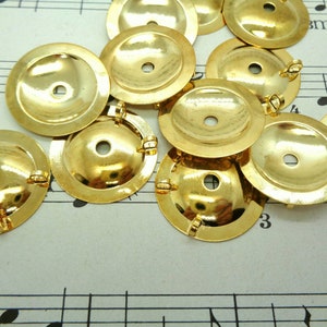 May include: A collection of shiny, gold-colored metal jewelry findings. Each piece is a small, shallow disc with a central hole and a loop for attaching to other components. The findings are scattered on a sheet of musical notation.
