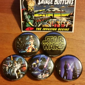 May include: Five Star Wars themed button pins. The pins feature different scenes from the original Star Wars movie. One pin has the text "Star Wars" in yellow letters on a black background. Another pin features a scene with Darth Vader and Luke Skywalker. The pins are displayed on a wooden surface.