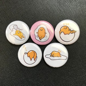 May include: Five round white and pink pinback buttons with cartoon egg yolk characters. Each button features a different illustration of the egg yolk character in various poses, with speech bubbles. The buttons are on a dark surface.