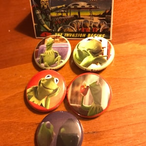 May include: Five round button pins featuring images of Kermit the Frog from the Muppets. The pins are on a wooden surface. The text "SAVAGE BUTTONS" and "THE INVASION BEGINS" are visible on a card in the background.