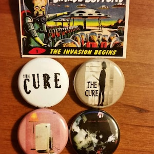 May include: An assortment of four round buttons and a vintage-style card. The buttons feature the band "The Cure" and various graphics. The card reads "Savage Buttons" and "The Invasion Begins" with an illustration of a Martian invasion.