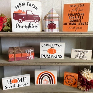 Op de afbeelding: Een verzameling houten borden met herfstthema's. De borden tonen pompoenen, vrachtwagens en zinnen als "Farm Fresh Pumpkins", "Happy Fall Y'all" en "Home Sweet Home". De kleuren zijn oranje, wit en bruin.