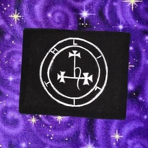 May include: A black square patch featuring a white sigil design. The sigil is a circular emblem with a cross-like symbol at the center, surrounded by other symbols. The background is a purple fabric with a starry pattern.