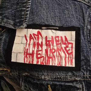 May include: A denim jacket with a rectangular patch. The patch features the text "I AM IN HELL, ME" in red, with a brick wall background. The patch is attached with a safety pin.