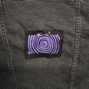 May include: Close-up of a dark gray denim jacket with a purple spiral patch. The patch is secured with a silver safety pin. The spiral design is painted on a black fabric patch.