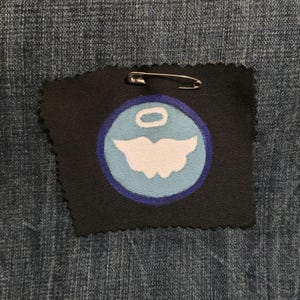 May include: A black fabric patch with a white and light blue design, featuring a stylized angel with a halo. A silver safety pin is attached to the patch. The patch is placed on a denim background.