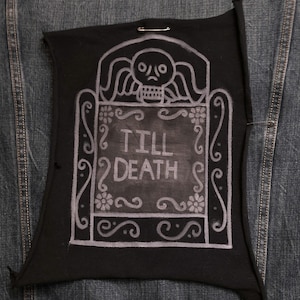 May include: Black fabric patch with a white tombstone design and the words "TILL DEATH". The tombstone features a skull, floral accents, and decorative scrollwork. The patch is pinned to a denim jacket.