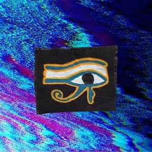 May include: A black square patch featuring the Eye of Horus symbol. The eye is outlined in gold with blue and white details, set against a vibrant, abstract blue and purple background.