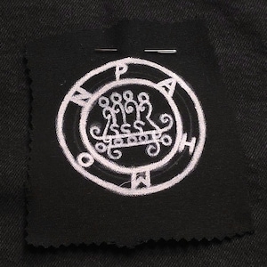 May include: A black fabric patch with a white embroidered sigil design. The sigil is circular with intricate symbols and letters. Two silver pins secure the patch to a dark background. The patch has a raw, unfinished edge.