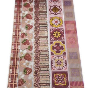 May include: A collection of decorative washi tape rolls with various patterns. Designs include strawberries, floral, brick, and geometric shapes in shades of pink, gold, and burgundy. The tape is ideal for crafts and scrapbooking.