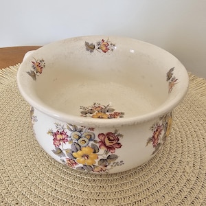 May include: Cream ceramic bowl featuring floral patterns in yellow, red, and blue. The bowl has a wide rim and a slightly textured surface. The floral design is repeated around the bowl's exterior. A vintage decorative piece.
