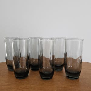 May include: A set of six clear drinking glasses with a smoky grey base. The glasses are cylindrical with a slightly flared rim. They are arranged on a wooden surface against a white background.