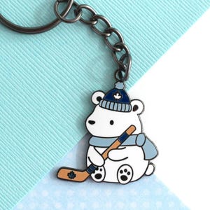 May include: A keychain with a white polar bear design. The bear wears a blue hat and scarf and holds a hockey stick. The hockey stick is brown and blue. The keychain is attached to a silver metal ring and chain. The bear has black eyes and paws.