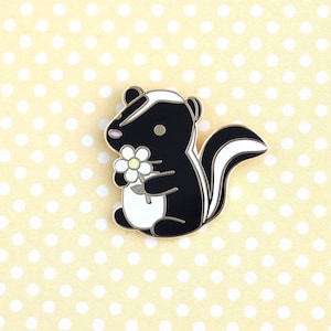 May include: A black and white skunk enamel pin, featuring a white flower with a yellow centre. The skunk has a white stripe and a pink nose. The pin is set against a yellow background with white polka dots.