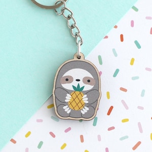 May include: A wooden keychain featuring a gray sloth holding a yellow pineapple. The keychain is attached to a silver metal ring and chain.
