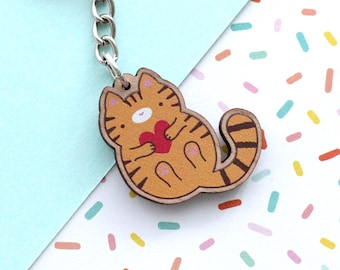 May include: A wooden keychain featuring a cartoon orange tabby cat holding a red heart. The keychain is attached to a silver metal ring.