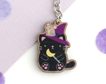 May include: A black cat wearing a purple witch hat with a small gray mouse on top. The cat is holding a yellow crescent moon on a stick. The cat is a wooden keychain.