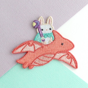May include: Embroidered patch showing a white rabbit in a teal cape, holding a purple wand, riding a coral dragon. The dragon has detailed wings. Set against a pastel background.