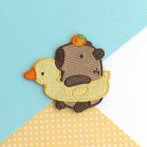 May include: Embroidered patch showing a brown bear with an orange fruit on its head, in a yellow duck-shaped float. The patch has a detailed, textured design, set against a blue and yellow background.