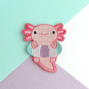 以下が含まれることがあります： ピンク色で、水色と紫の浮き輪を着けた、笑顔のウーパールーパーの刺繍ワッペン。ウーパールーパーは詳細なエラとフレンドリーな表情をしています。このワッペンは、衣類やアクセサリーに縫い付けたり、アイロンで接着したりできます。