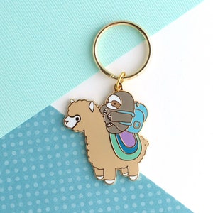 Gold plated keychain of little grey sloths wearing a backpack and riding on top of a beige alpaca (or llama)