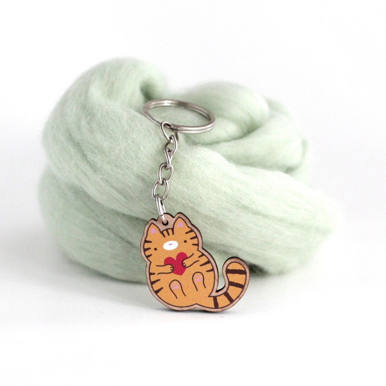 May include: A wooden keychain featuring a cartoon orange cat holding a red heart. The keychain is attached to a silver key ring.