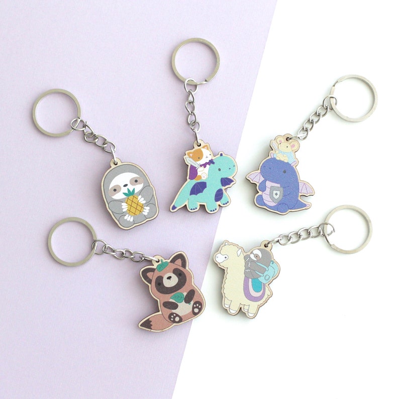 May include: Six colorful wooden keychains featuring various animal characters. The keychains include a sloth holding a pineapple, a dragon with a cat riding on its back, a llama with a sloth on its back, a raccoon holding a leaf, a dragon with a mouse riding on its back, and a sloth holding a book.
