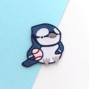 Op de afbeelding: Geborduurde patch met een cartoon blauwe gaai die een honkbal vasthoudt. De patch heeft een marineblauwe omtrek, een witte body en grijze accenten. De honkbal is rood en wit. Ideaal voor jassen, tassen of andere stoffen items.