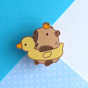 Peut inclure: Un ours brun avec une orange sur la tête est assis sur un flotteur de canard en caoutchouc jaune. L'ours porte une chemise à rayures brunes et blanches. Le flotteur de canard a un bec jaune et des pieds orange.