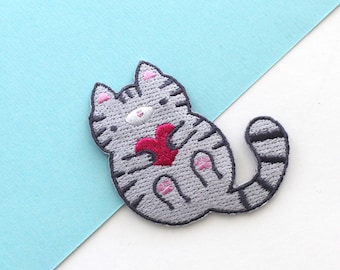 May include: Gray embroidered iron-on patch of a cat holding a red heart. The cat has pink ears, nose, and paws.