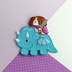 Könnte beinhalten: Ein gestickter Aufnäher mit einem blauen Triceratops, auf dem ein braunes und weißes Hündchen reitet. Der Hund trägt einen lila-weißen Schal und hält ein Schwert.