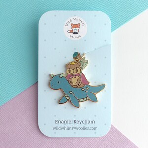 May include: Enamel keychain featuring a cartoon bear wearing a crown and cape riding a blue sea monster. The keychain is attached to a light blue card with the text "Enamel Keychain" and "wildwhimsywoolies.com". The card has a fox logo.