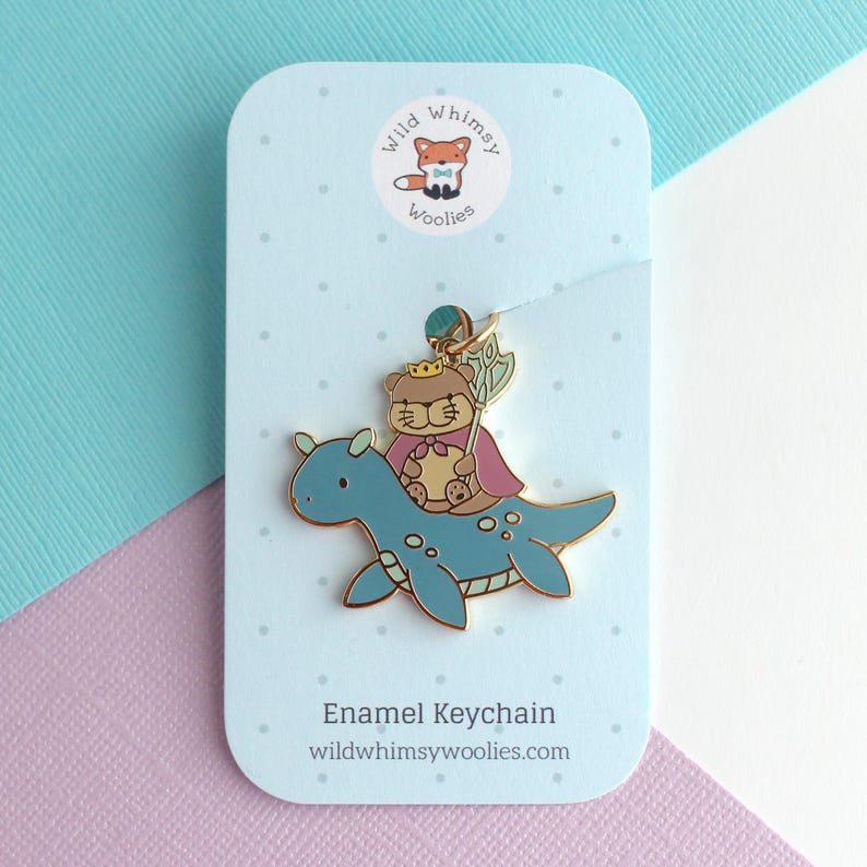 May include: Enamel keychain featuring a cartoon bear wearing a crown and cape riding a blue sea monster. The keychain is attached to a light blue card with the text "Enamel Keychain" and "wildwhimsywoolies.com". The card has a fox logo.