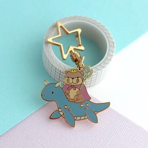 May include: A gold-colored metal keychain featuring a cartoon bear wearing a crown and cape, riding a blue dinosaur-like creature. The keychain has a star-shaped clasp and is set against a pastel background.