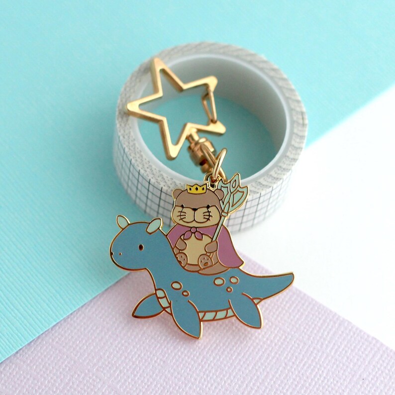 May include: A gold-colored metal keychain featuring a cartoon bear wearing a crown and cape, riding a blue dinosaur-like creature. The keychain has a star-shaped clasp and is set against a pastel background.