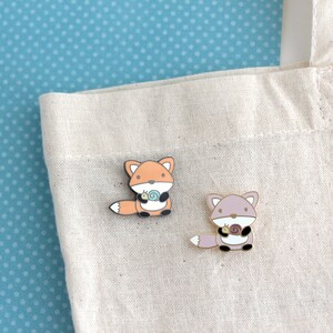 May include: Two enamel pins featuring cartoon foxes holding snails. The fox on the left is orange and the fox on the right is purple.