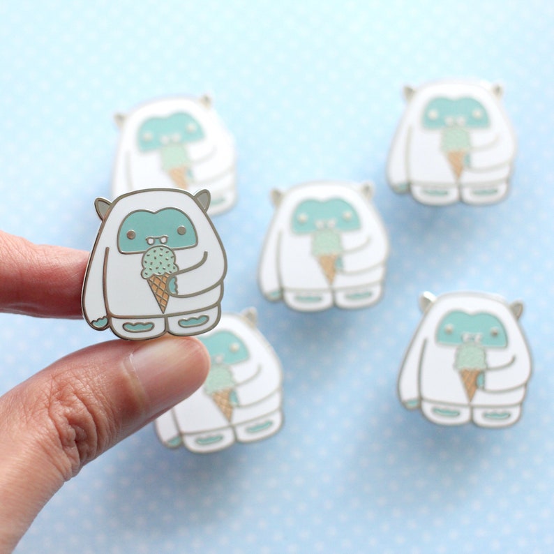 Hand showing an enamel pin of a white and teal yeti enjoying a mint chocolate chip ice cream
