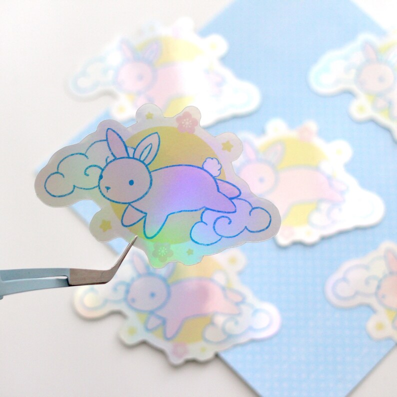 May include: A set of iridescent holographic stickers featuring a cartoon bunny rabbit flying on a cloud with a moon and stars in the background. The stickers are perfect for adding a touch of whimsy to laptops, water bottles, journals, and more.
