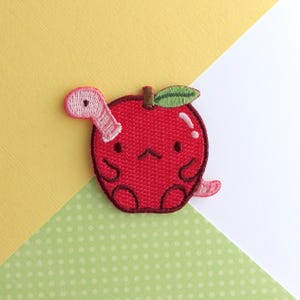 May include: Embroidered patch featuring a red apple with a cute, sad face and a pink worm emerging from it. The apple has a green leaf and a brown stem. The patch is on a background of yellow, white, and green polka dot paper.