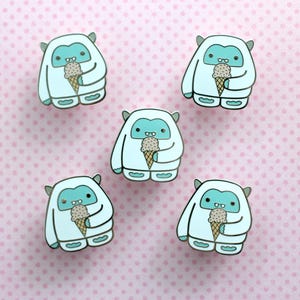 May include: Five white Yeti pins with blue accents, each holding a brown and white ice cream cone. The pins are on a pink background with white polka dots.